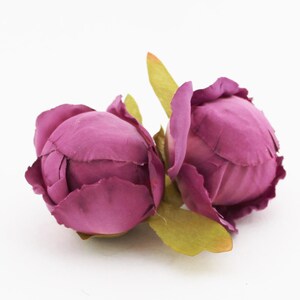 May include: Two artificial peony flower heads in a deep magenta hue. The petals are tightly furled, with a textured appearance. Green leaves are visible at the base of the blooms, set against a white background. These faux flowers are suitable for crafting and decor.