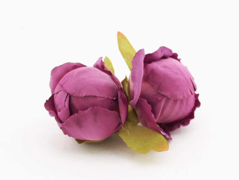 May include: Two artificial peony flower heads in a deep magenta hue. The petals are tightly furled, with a textured appearance. Green leaves are visible at the base of the blooms, set against a white background. These faux flowers are suitable for crafting and decor.