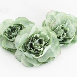 May include: Three sage green artificial peony flowers with multiple layers of petals. The flowers have a soft, delicate appearance and are arranged on a white background. The edges of some petals have a hint of pink.