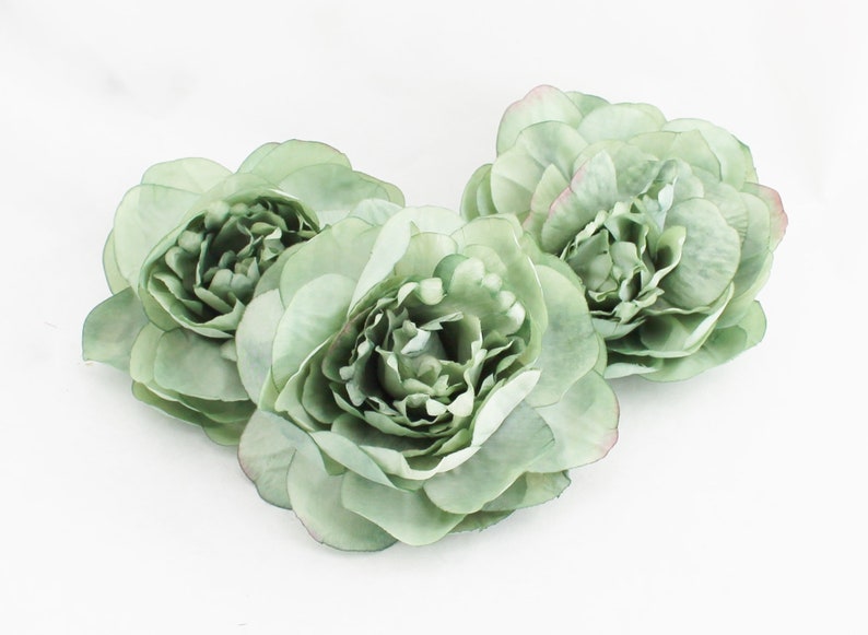 May include: Three sage green artificial peony flowers with multiple layers of petals. The flowers have a soft, delicate appearance and are arranged on a white background. The edges of some petals have a hint of pink.