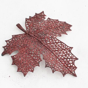 May include: A decorative, glittery, burgundy-colored leaf ornament. The leaf has a detailed, openwork design with a network of small triangles. The stem is a darker shade of burgundy. This item is suitable for fall and holiday decor.