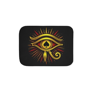 May include: A rectangular black mat featuring a central graphic of the Eye of Horus in gold and red. The pupil of the eye is replaced by a black spade symbol. The design includes radiating lines and dots, creating a sunburst effect around the eye.