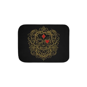 May include: A rectangular black mat featuring a central gold-colored skull illustration surrounded by intricate floral vines and thorns. The skull incorporates red playing card suit symbols, specifically a diamond on the forehead and a heart for an eye.