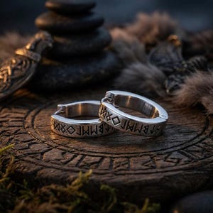 Op de afbeelding: Een paar zilverkleurige oorringen met gegraveerde zwarte runensymbolen op de buitenkant. De oorbellen hebben een scharniersluiting en een gepolijste metalen afwerking, voorzien van gedetailleerde, in het metaal geëtste tekens in oude stijl.