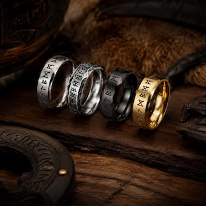 Op de afbeelding: Vier metalen ringen op een rij, elk voorzien van gegraveerde runensymbolen. De eerste ring is van gepolijst zilver, de tweede van getextureerd zilver, de derde van matzwart en de vierde van gepolijst goudkleurig metaal.