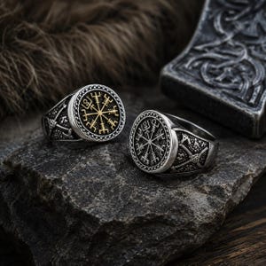 Peut inclure: Deux bagues argentées présentant une face circulaire ornée du symbole Vegvisir. L'une des bagues possède un centre doré, tandis que l'autre est entièrement argentée. Les deux anneaux présentent des gravures latérales de haches croisées.