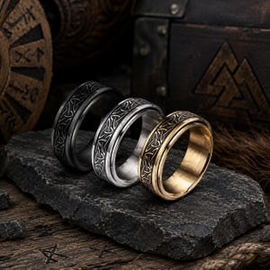 May include: Three metal rings arranged in a row, each featuring a continuous engraved Celtic triquetra knot pattern. The rings are finished in matte black, polished silver, and brushed gold tones, showcasing a wide band design with smooth edges.