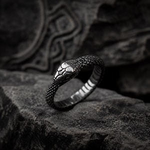 Peut inclure: Une bague en métal argenté foncé en forme de serpent enroulé. L'anneau présente un motif texturé rappelant des écailles sur toute sa surface, avec une tête de serpent détaillée et lisse au centre. L'intérieur de l'anneau est strié.