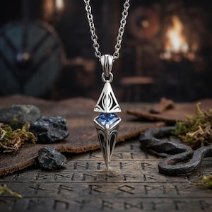 May include: A silver-toned metal pendant necklace featuring a geometric, pointed design with an intricate, dark-etched pattern and a central, faceted blue gemstone. The pendant hangs from a matching silver-toned chain link necklace.