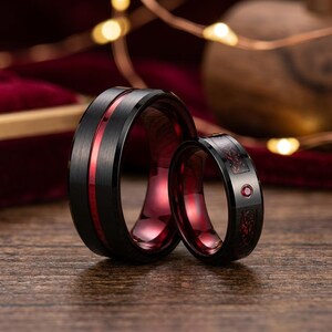 Peut inclure: Deux bagues noires avec une finition brossée. La plus grande bague présente une fine incrustation rouge centrée. La plus petite bague affiche une incrustation à motif rouge et une petite pierre précieuse rouge circulaire sertie.