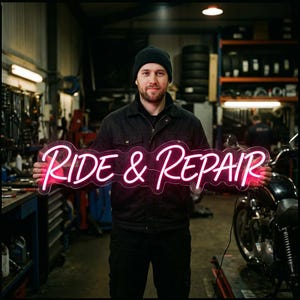 May include: A bright pink neon sign featuring the text "RIDE & REPAIR" in a cursive script font. The sign is illuminated and displays a glowing, vibrant pink light, showcasing the words clearly against the dark background of a workshop.