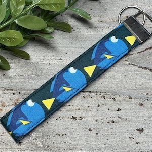 May include: A fabric keychain wristlet featuring a repeating pattern of blue fish with yellow fins on a dark green background. The strap is secured with a silver-toned metal hardware clamp and a circular split ring for attaching keys.