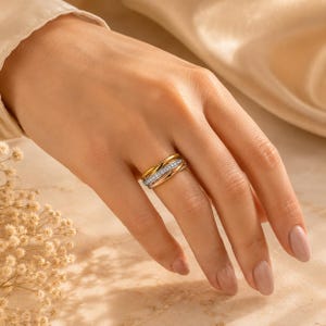 Pode incluir: Um anel de várias bandas com três aros entrelaçados em ouro amarelo, rosa e branco. A banda central de ouro branco é cravejada com uma fila de pequenas pedras claras de corte redondo que brilham na superfície do anel, criando um visual.