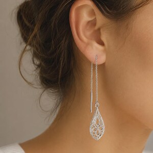 May include: A silver-toned threader earring featuring a delicate, teardrop-shaped filigree pendant. The earring consists of a thin chain that passes through the earlobe, ending in a straight metal post on one side and the ornate, openwork charm.