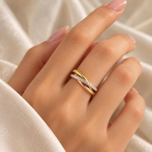 May include: A tri-color crossover ring featuring three intertwined bands in yellow gold, rose gold, and a silver-toned metal band encrusted with a row of small, clear, round-cut stones that catch the light across the surface of the band.