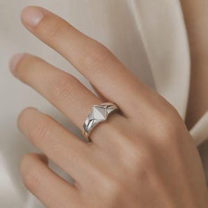 以下が含まれることがあります： 多面的な幾何学模様のデザインが施されたシルバー調のリング。中央には尖った三角形のパーツがあしらわれています。バンドは滑らかで光沢のある仕上げで、指の裏側に向かってわずかに細くなる構造的で角張った形状をしています。