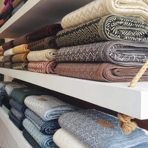 Puede incluir: Una colección de mantas tejidas dobladas apiladas en estantes blancos. Las mantas presentan varios patrones geométricos, incluidos zigzags y diamantes, en una paleta de crema, marrón, gris, azul y verde, con pequeñas etiquetas rectangulares.