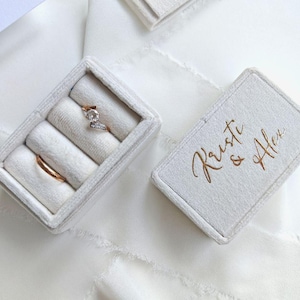 May include: A rectangular white velvet ring box with a removable lid featuring the gold script text Kristi & Alex. The open box contains two ring slots, one holding a silver-toned ring with a clear stone and the other a plain gold band.