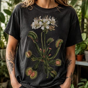 May include: A charcoal black short-sleeve t-shirt featuring a detailed botanical illustration of a Venus flytrap plant with green leaves, spiky traps, and white flowers centered on the front of the garment. The fabric has a faded, washed-out texture.