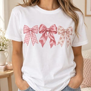 May include: A white short-sleeved t-shirt featuring a graphic print of three distinct pink bows across the chest. The left bow has a pink and white gingham pattern, the centre bow is solid pink, and the right bow has a pink floral pattern.