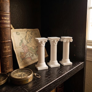 May include: Three white miniature architectural columns featuring fluted shafts and detailed capitals. Each column stands on a square base, showcasing classical design elements. These small decorative pillars are arranged in a row on a dark surface.