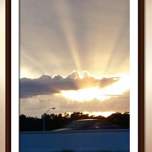 May include: A framed photograph of a sunset with bright golden light rays beaming upward through dark clouds above a silhouette of trees and a blurred vehicle moving along a road. The frame is dark brown with a thick white mat border.