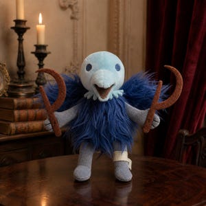 May include: A plush bird-like creature with a light blue head, a dark blue shaggy body, and grey limbs. It features a small white bandage on its left leg and holds two brown, curved, sickle-shaped felt objects in its hands, one in each hand.
