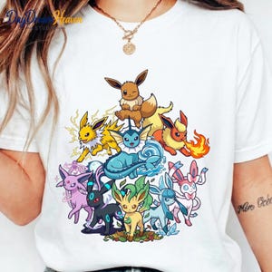 May include: A white t-shirt featuring a colorful graphic print of nine different Eevee evolution characters arranged in a cluster. The design includes Eevee, Jolteon, Vaporeon, Flareon, Espeon, Umbreon, Leafeon, Glaceon, and Sylveon with elemental effects.