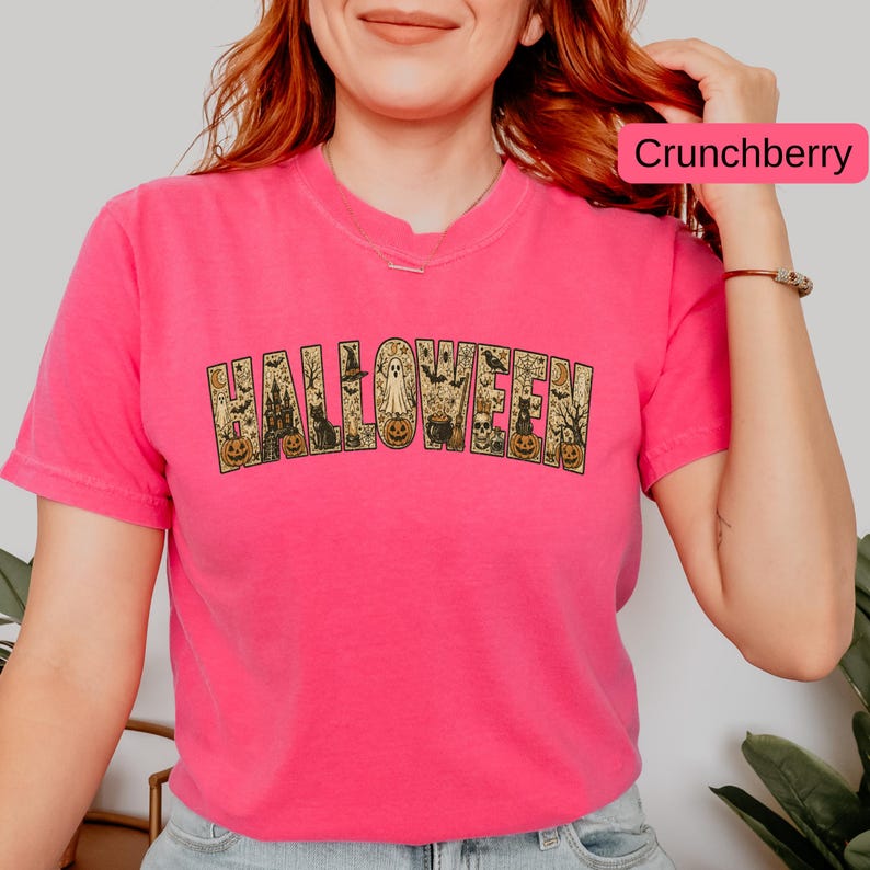 May include: A bright pink short-sleeve t-shirt featuring the word HALLOWEEN printed in a large, arched font. The letters are filled with detailed illustrations of pumpkins, ghosts, black cats, bats, skulls, and haunted houses in gold.