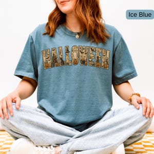 May include: An ice blue short-sleeve t-shirt featuring the word HALLOWEEN printed in large block letters. The letters contain detailed illustrations of pumpkins, a haunted house, a black cat, a ghost, a skull, and bats in shades of orange and black.