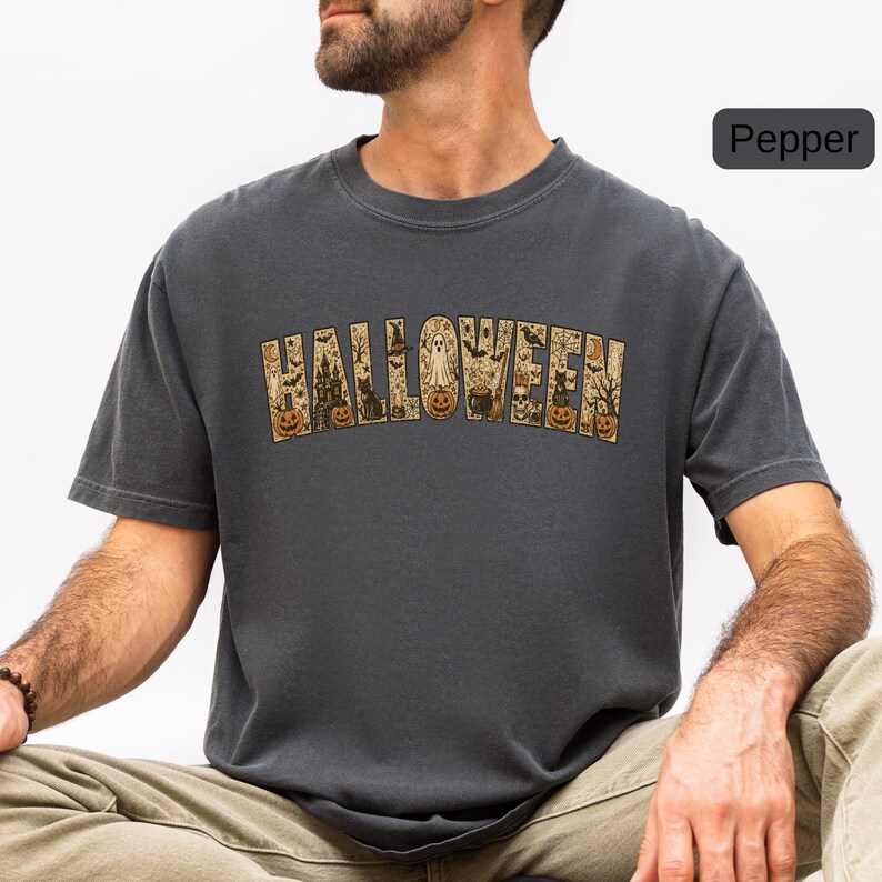 May include: A charcoal gray short-sleeve t-shirt featuring the word HALLOWEEN printed in a large, arched font. The letters are filled with intricate illustrations of pumpkins, ghosts, black cats, skulls, bats, and a haunted house.
