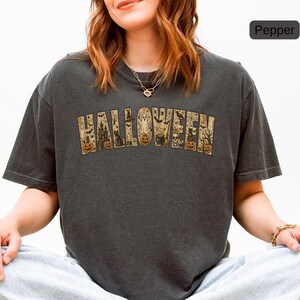 May include: A charcoal gray short-sleeve t-shirt featuring the word HALLOWEEN printed in large block letters. Each letter contains detailed illustrations of pumpkins, a ghost, a haunted house, a black cat, a cauldron, and bats.