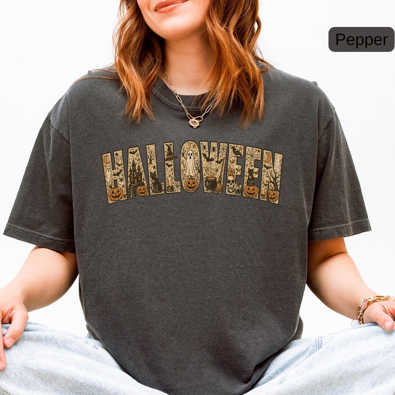 May include: A charcoal gray short-sleeve t-shirt featuring the word HALLOWEEN printed in large block letters. Each letter contains detailed illustrations of pumpkins, a ghost, a haunted house, a black cat, a cauldron, and bats.