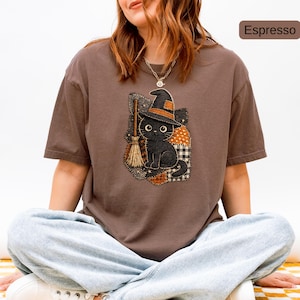 May include: An espresso-coloured short-sleeve t-shirt featuring a graphic of a black cat wearing a witch hat next to a broomstick. The design is set against a patchwork background of orange, black, and white patterns, including polka dots and plaid.