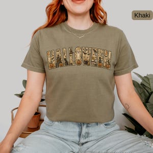 May include: A khaki crew neck t-shirt featuring the word HALLOWEEN printed in large block letters. The letters are filled with detailed illustrations of pumpkins, ghosts, a haunted house, black cats, bats, spiders, and a cauldron.