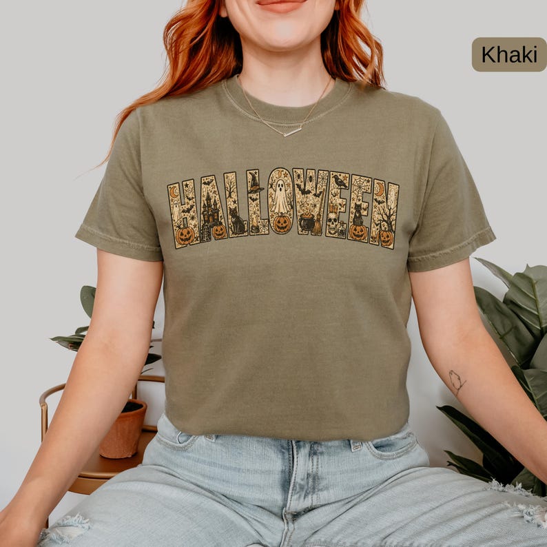 May include: A khaki crew neck t-shirt featuring the word HALLOWEEN printed in large block letters. The letters are filled with detailed illustrations of pumpkins, ghosts, a haunted house, black cats, bats, spiders, and a cauldron.