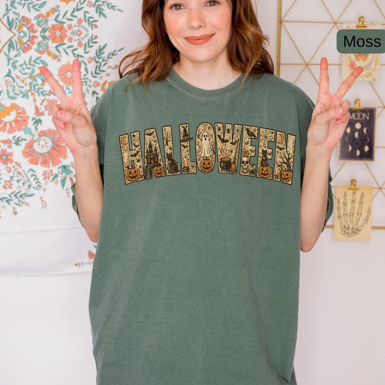 May include: A moss green short-sleeve t-shirt featuring the word HALLOWEEN printed in large block letters. Each letter contains detailed illustrations of pumpkins, a haunted house, a ghost, a black cat, a skull, a cauldron, and spider webs.
