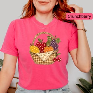 Op de afbeelding: Een felroze T-shirt met korte mouwen en een centrale afbeelding van een gevlochten mand gevuld met diverse vruchten, waaronder een gele peer, een rode appel, een groen gepatroonneerde peer, een paarse tros druiven en rode kersen.