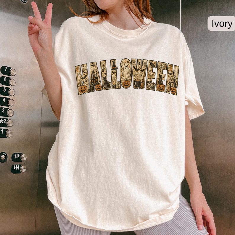 May include: An ivory short-sleeve t-shirt featuring the word HALLOWEEN printed in large block letters. Each letter contains detailed illustrations of pumpkins, ghosts, bats, a haunted house, a black cat, a cauldron, and a spider web.