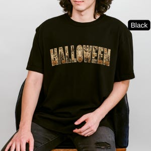 May include: A black short-sleeved t-shirt featuring the word HALLOWEEN printed in a large, arched font. The letters are filled with a detailed graphic pattern of pumpkins, ghosts, black cats, bats, a haunted house, and a cauldron.