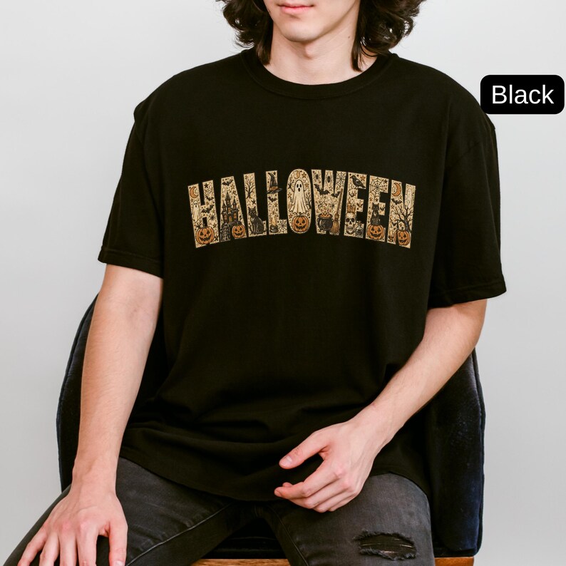 May include: A black short-sleeved t-shirt featuring the word HALLOWEEN printed in a large, arched font. The letters are filled with a detailed graphic pattern of pumpkins, ghosts, black cats, bats, a haunted house, and a cauldron.