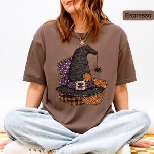 Puede incluir: Una camiseta de manga corta color espresso con un gráfico de un sombrero de bruja negro con banda morada, hebilla plateada y una araña colgante. El diseño incluye patrones de patchwork en morado, naranja y cuadros y lunares negros.