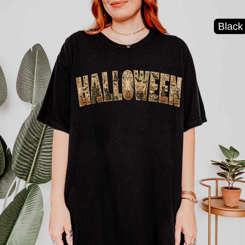 May include: A black t-shirt featuring the word HALLOWEEN printed in a large, arched font. The letters are filled with a detailed, tan-colored graphic pattern depicting various spooky elements including ghosts, pumpkins, cats, and a castle.