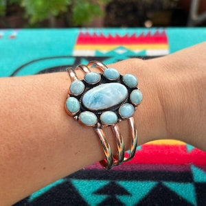 May include: A copper cuff bracelet featuring a large oval central light blue stone surrounded by eight smaller oval light blue stones. The metal band consists of three parallel wires that meet at the sides to support the stone cluster setting.