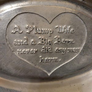 May include: An oval-shaped silver-toned metal tray featuring a central heart-shaped indentation. Embossed inside the heart is the text: A Plump Wife and a Big Barn never did any man harm. The metal surface has a brushed, matte finish throughout.