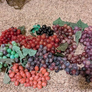 Op de afbeelding: Een verzameling kunstmatige druiventrossen in diverse kleuren, waaronder lichtgroen, donkerrood, dieppaars en zwart. Elke tros bevat realistische plastic fruitbolletjes bevestigd aan steeltjes met groene stoffen bladeren ertussen.