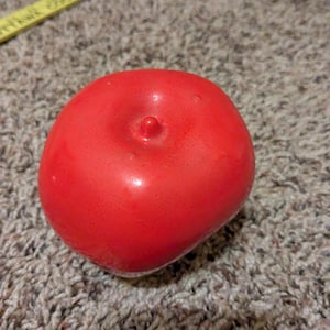 May include: A bright red, smooth, round apple-shaped object with a small central stem nub. A yellow ruler is visible in the upper portion of the frame, showing a portion of the measurement scale in inches along the top edge of the object.