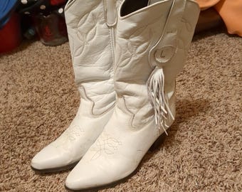 May include: A pair of white leather cowboy boots featuring a pointed toe, mid-calf height, and decorative stitching on the shaft. Each boot includes a circular leather patch with a fringe detail hanging from the side pull tab.