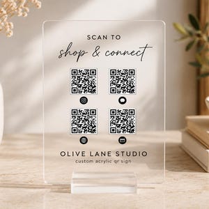 May include: A clear rectangular acrylic sign with a stand featuring the text SCAN TO shop & connect at the top, four black and white QR codes in the center with small social media icons below them, and OLIVE LANE STUDIO custom acrylic qr sign at the bottom.