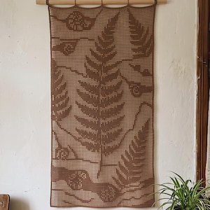 May include: A rectangular brown crochet wall hanging featuring a detailed fern leaf pattern. The textile is suspended from a horizontal wooden dowel with a thin cord attached for mounting, displaying a grid-like texture throughout the design.
